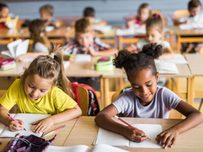 Happy Asian and African American elementary students writing in their notebooks while having a class at school. Their classmates are in the background.