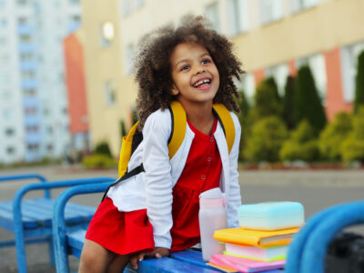 schoolgirl eating outdoors next the school. Healthy school breakfast for child. Food for lunch, lunchboxes with sandwiches, fruits, vegetables, and water.