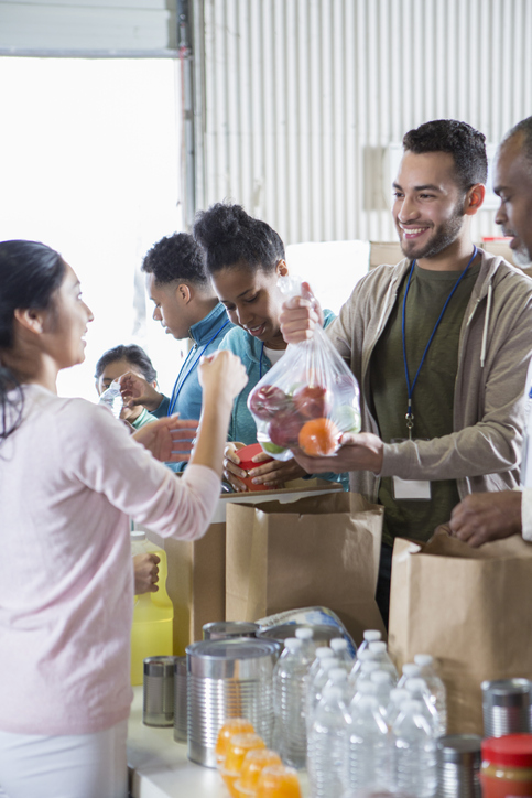 A group of food bank volunteers stand at a table in a warehouse and organize food. A young man accepts a donation of a bag of apples.