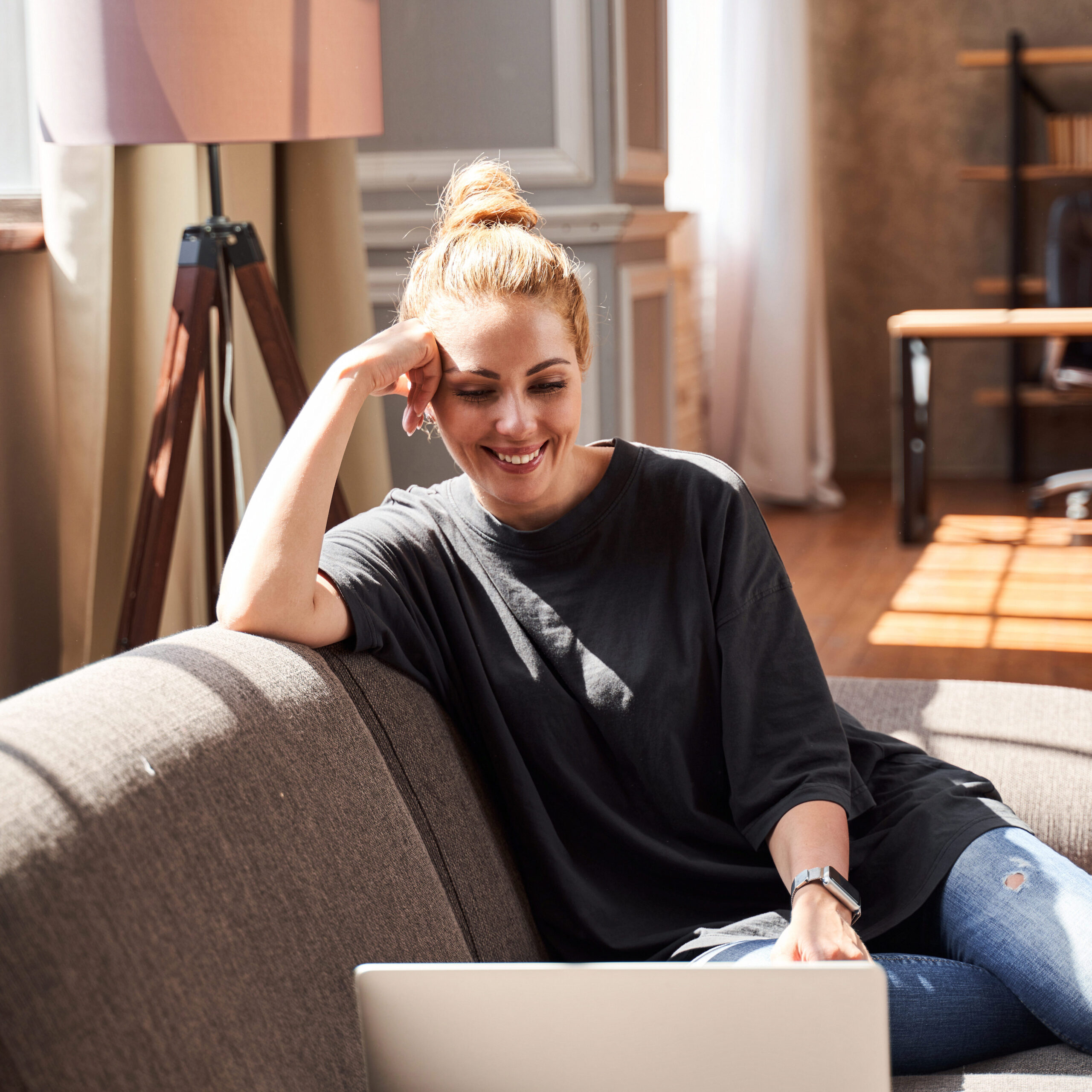 Cheerful young lady in jeans and T-shirt sitting on the sofa and smiling to the laptop camera during the video call