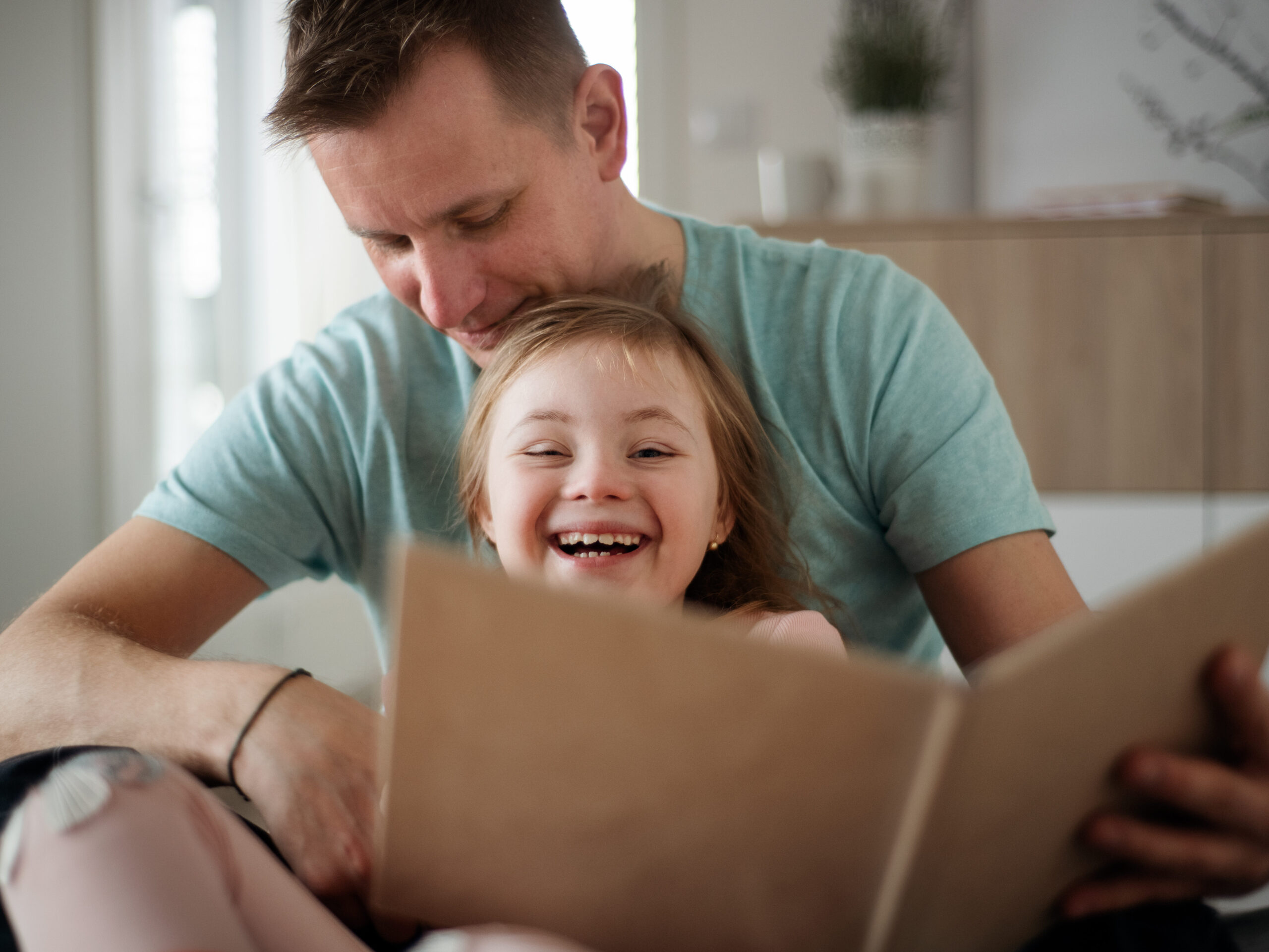 A father reading book to his little daughter with Down syndrome when sitting on bed at home.