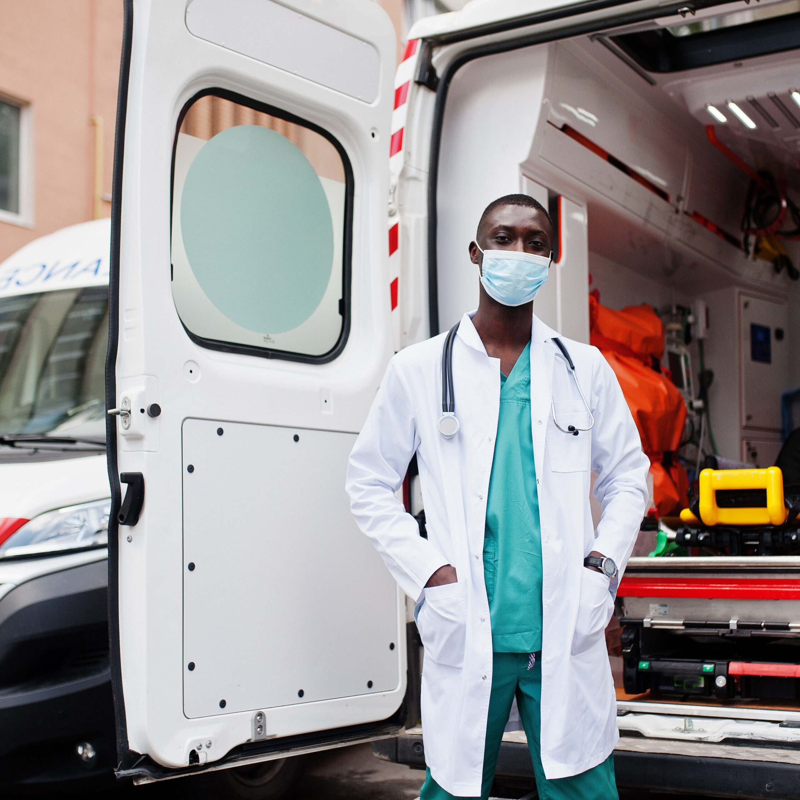 African male paramedic in face protective medical mask standing in front of ambulance car.