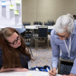 two woman lean on table, writing on post it notes